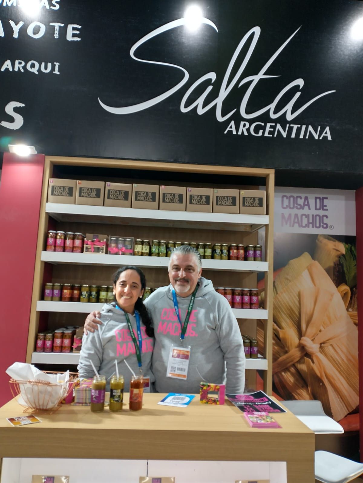 Stand de Salta, Argentina, con dos personas detrás de una mesa llena de frascos y botellas en una feria comercial.