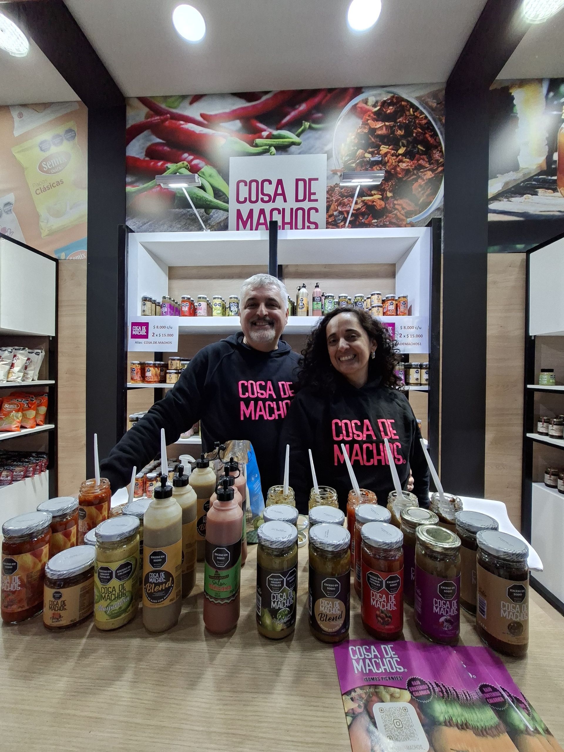 Dos personas detrás de un puesto de mercado exhiben frascos y botellas de salsas con la etiqueta “Casa de Máquinas”.