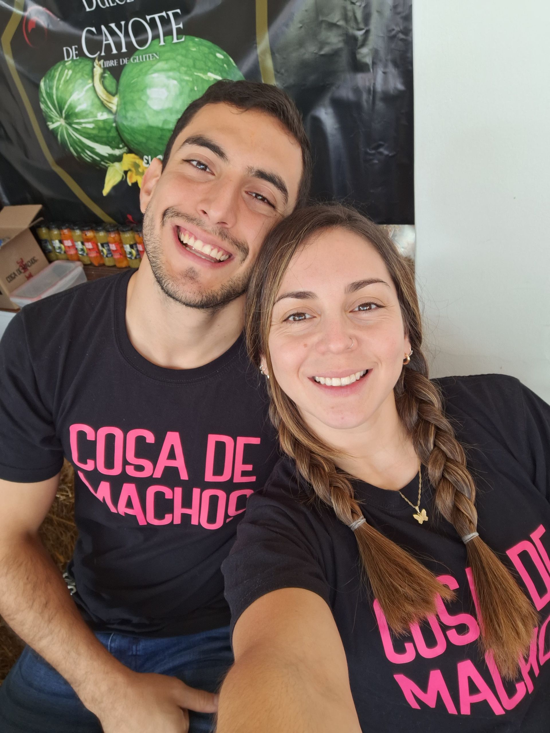 Pareja sonriente con camisetas negras con texto rosa, posando para una selfie en el interior.