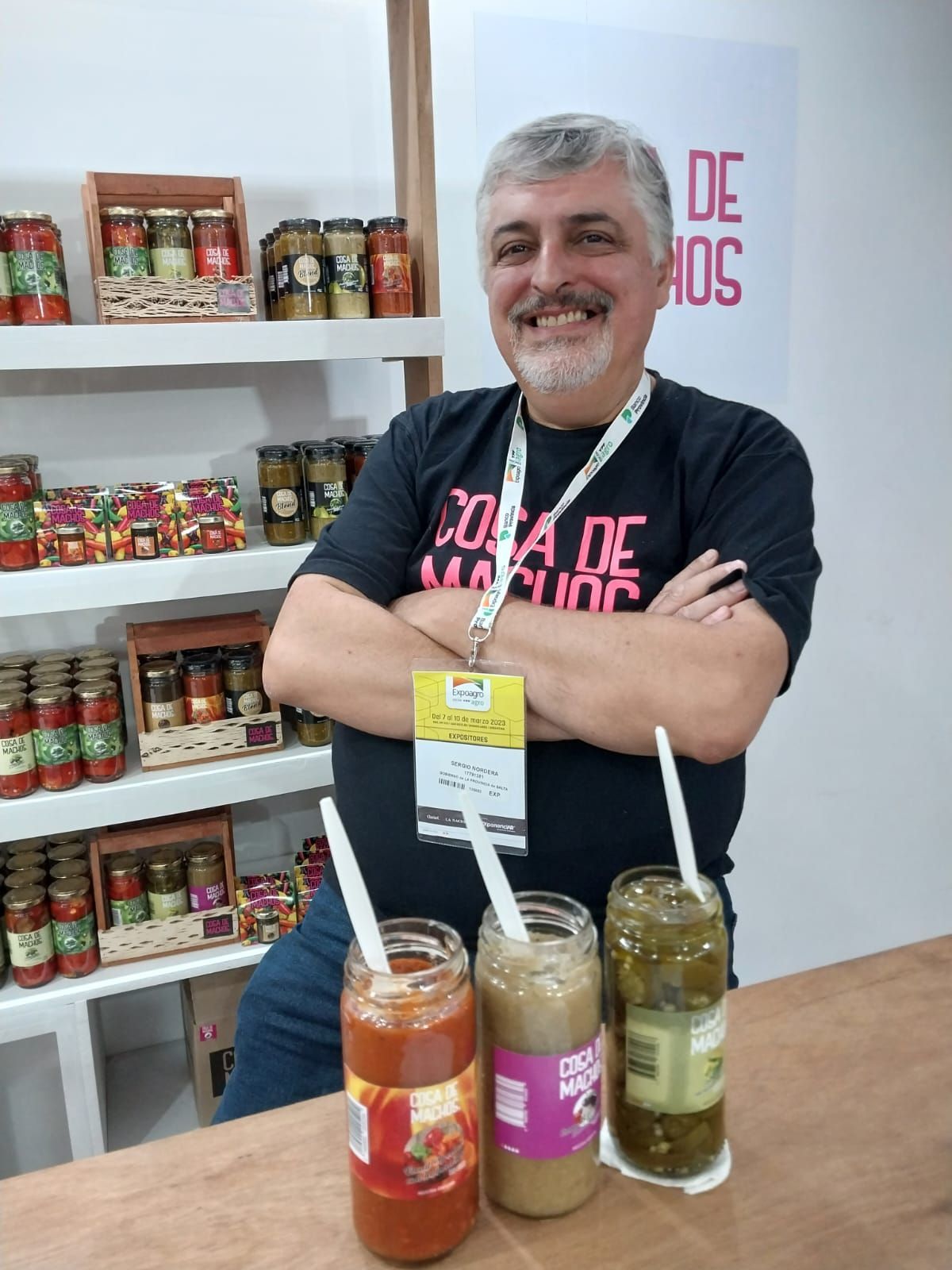 Un hombre sonríe detrás de una exhibición de frascos y frascos de mermelada con cucharas sobre una mesa.