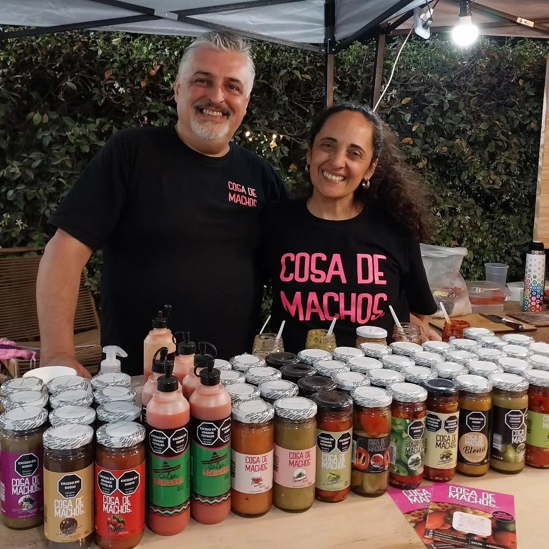 Dos vendedores sonríen detrás de un puesto de mercado repleto de tarros y botellas de salsas y conservas.