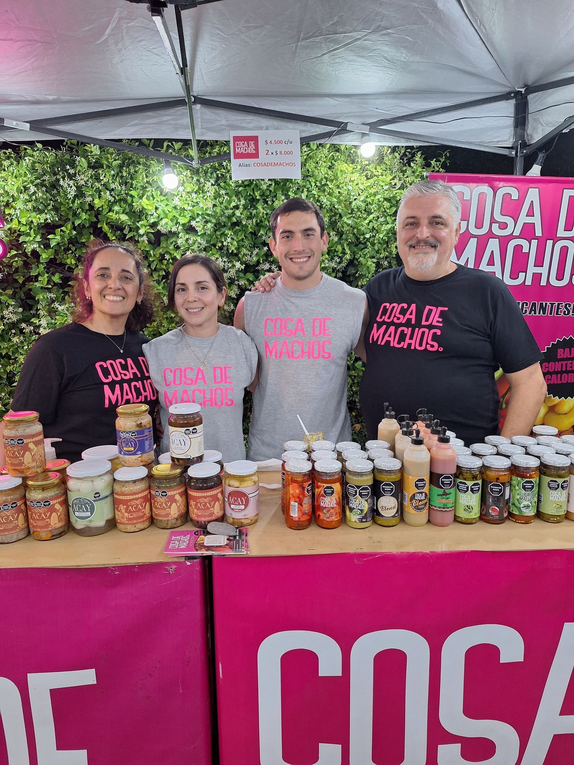 Cuatro personas detrás de una mesa llena de salsas embotelladas en un puesto de mercado con una pancarta de color rosa