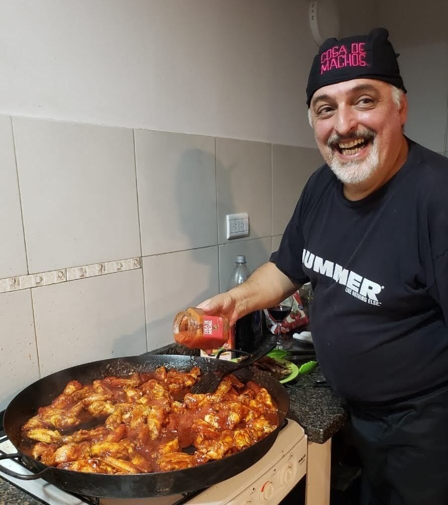 Un hombre sonriente, con sombrero y camisa negros, está de pie junto a una gran sartén con salsa de tomate 