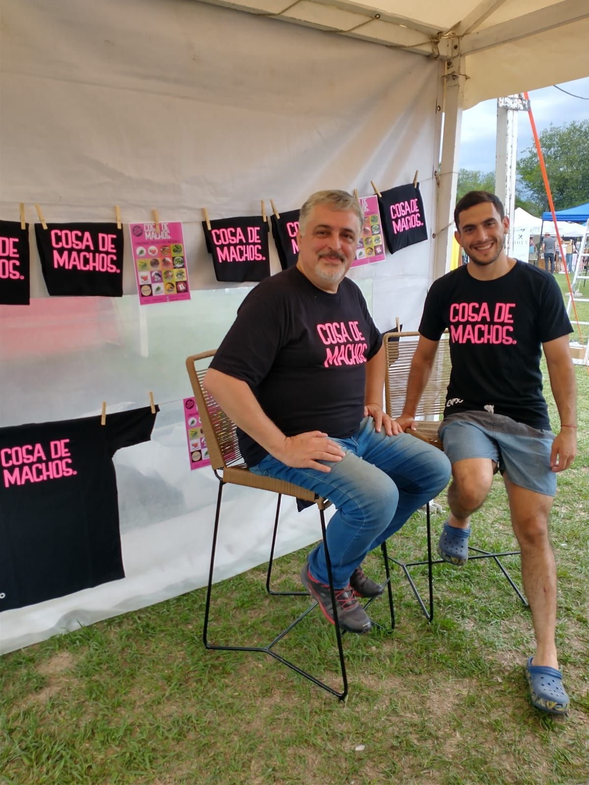 Dos hombres posando en una carpa con camisetas negras de “Cica de Madre” colgando detrás de ellos.