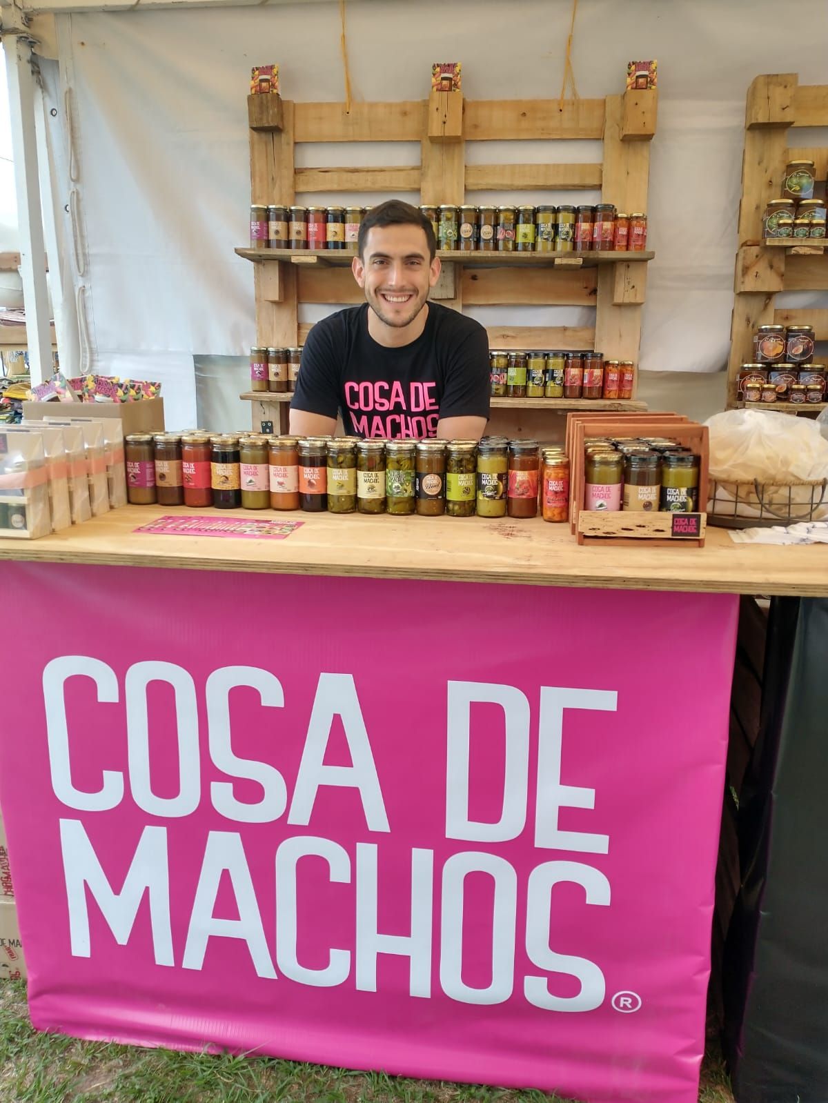 Vendedor sonriente detrás de una mesa llena de frascos en el puesto de Cosa de Machos con una pancarta rosa brillante.