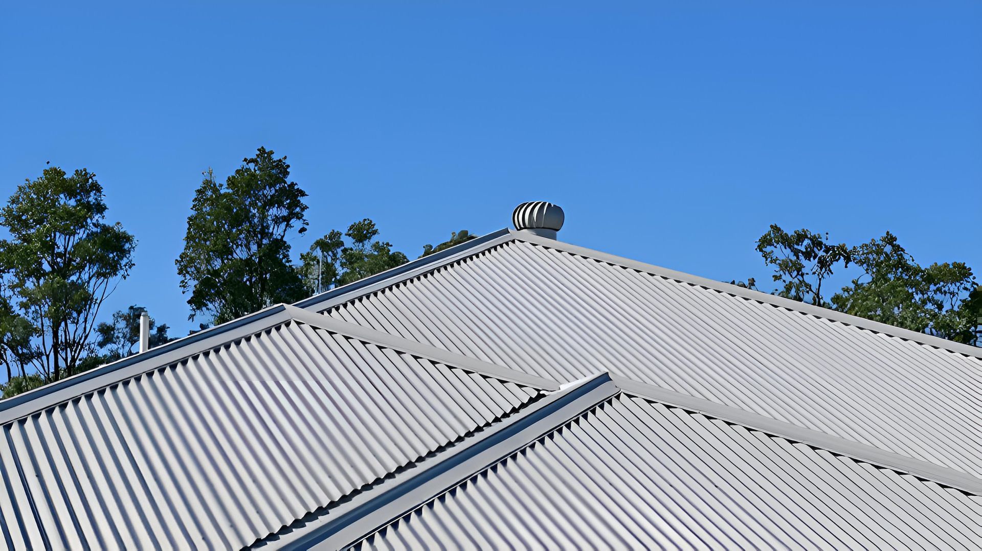 Grey corrugated metal roof with ridges and a rooftop ventilator set against a clear blue sky