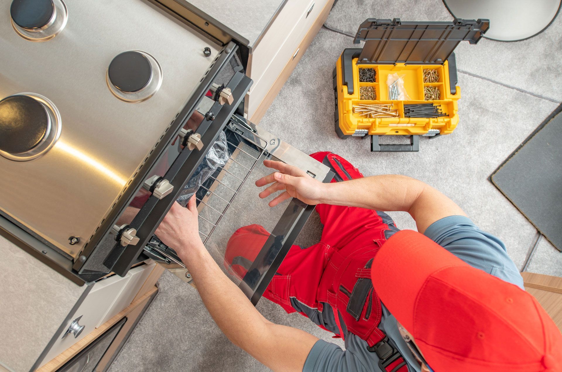 Person repairing a gas stove, wearing a red hat, with a yellow toolbox nearby.