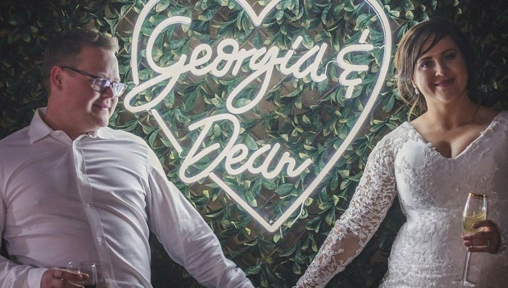 A bride and groom are holding hands in front of a neon sign that says `` georgia 's dear ''.