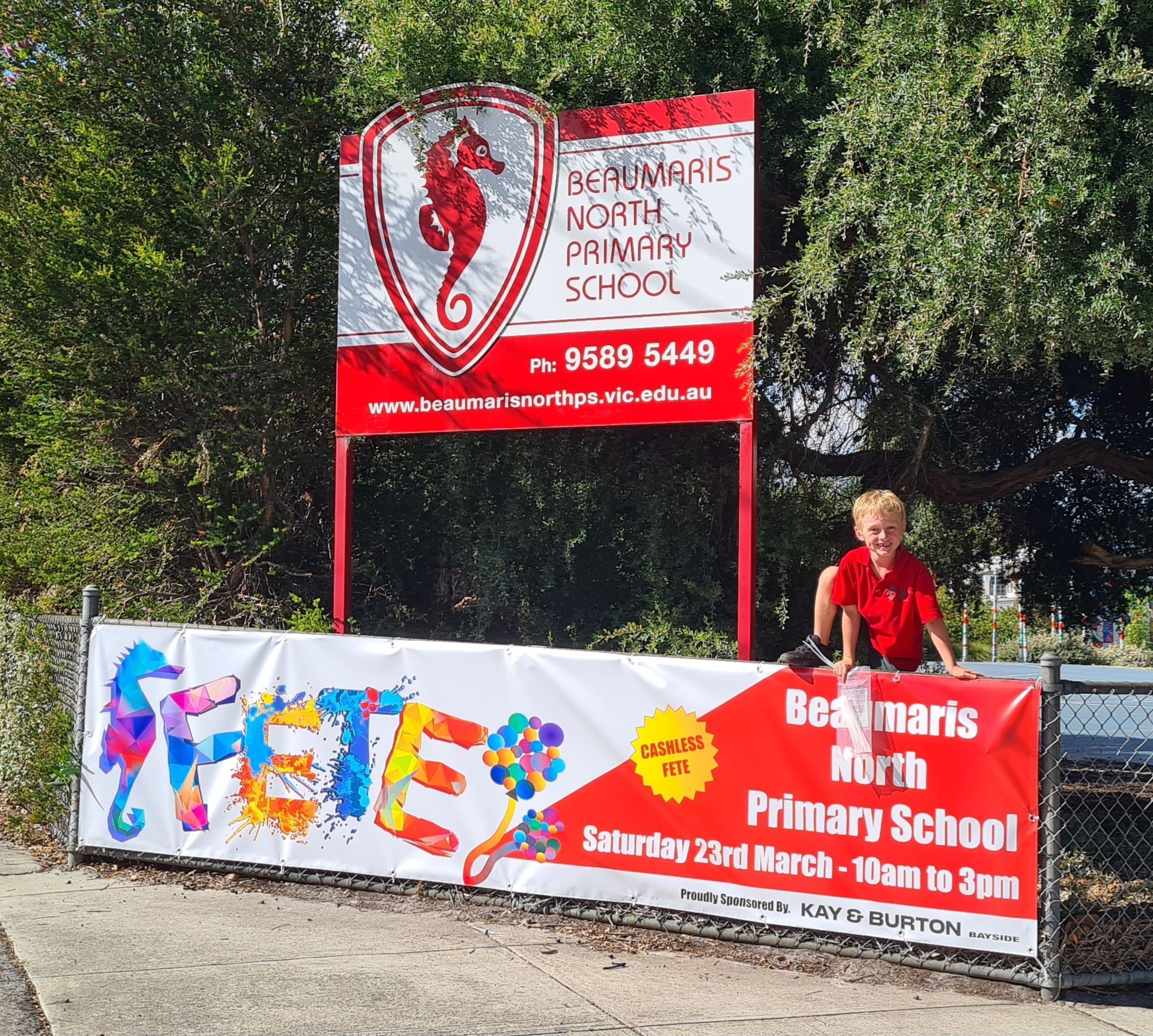 A boy in a red shirt stands in front of a sign for bernamaris north primary school