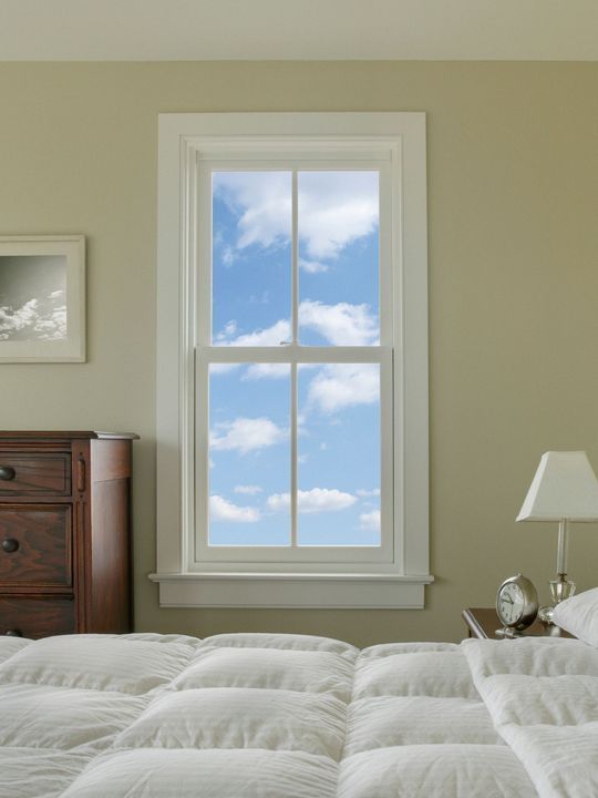 Camera da letto con finestra con cornice bianca che mostra un cielo azzurro con nuvole; letto e comodino visibili.