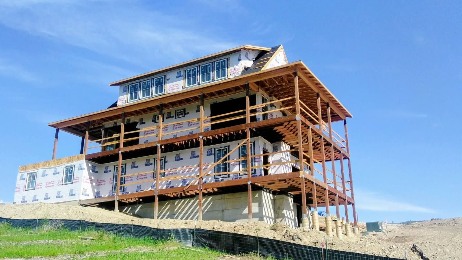 A multi-story house under construction with exposed wooden framing and white weather-resistant barriers, set against a hill.