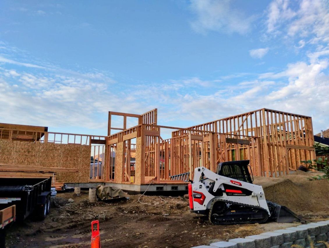 A wooden frame construction of a new building under a blue sky, with a white skid steer loader parked in the foreground.
