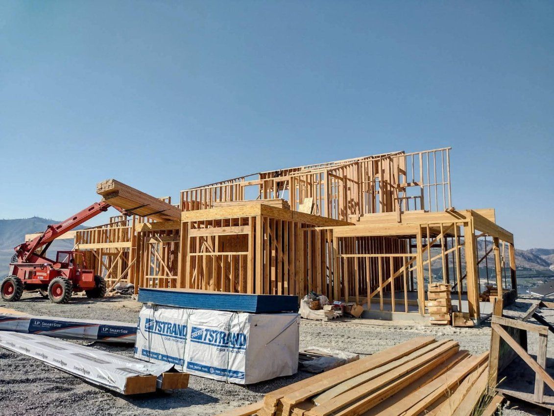 A red construction vehicle lifts a large wooden beam onto the wood-framed structure of a house under construction.