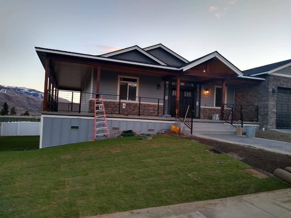 A modern, single-story house with a wrap-around porch, stone accents, and a sloped gray roof at twilight.
