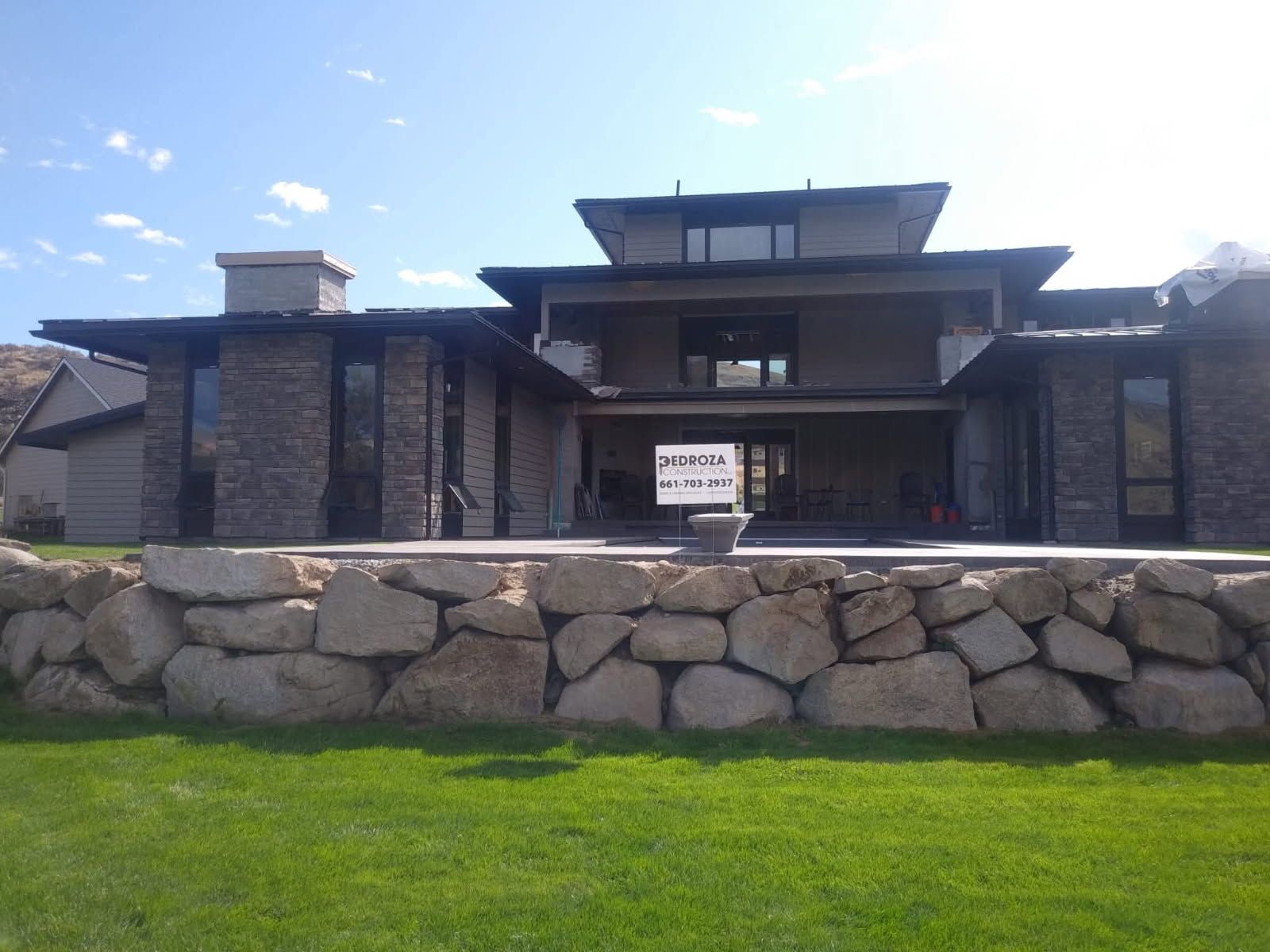 A modern, two-story house with stone pillars and a large, tiered rock retaining wall in the foreground.