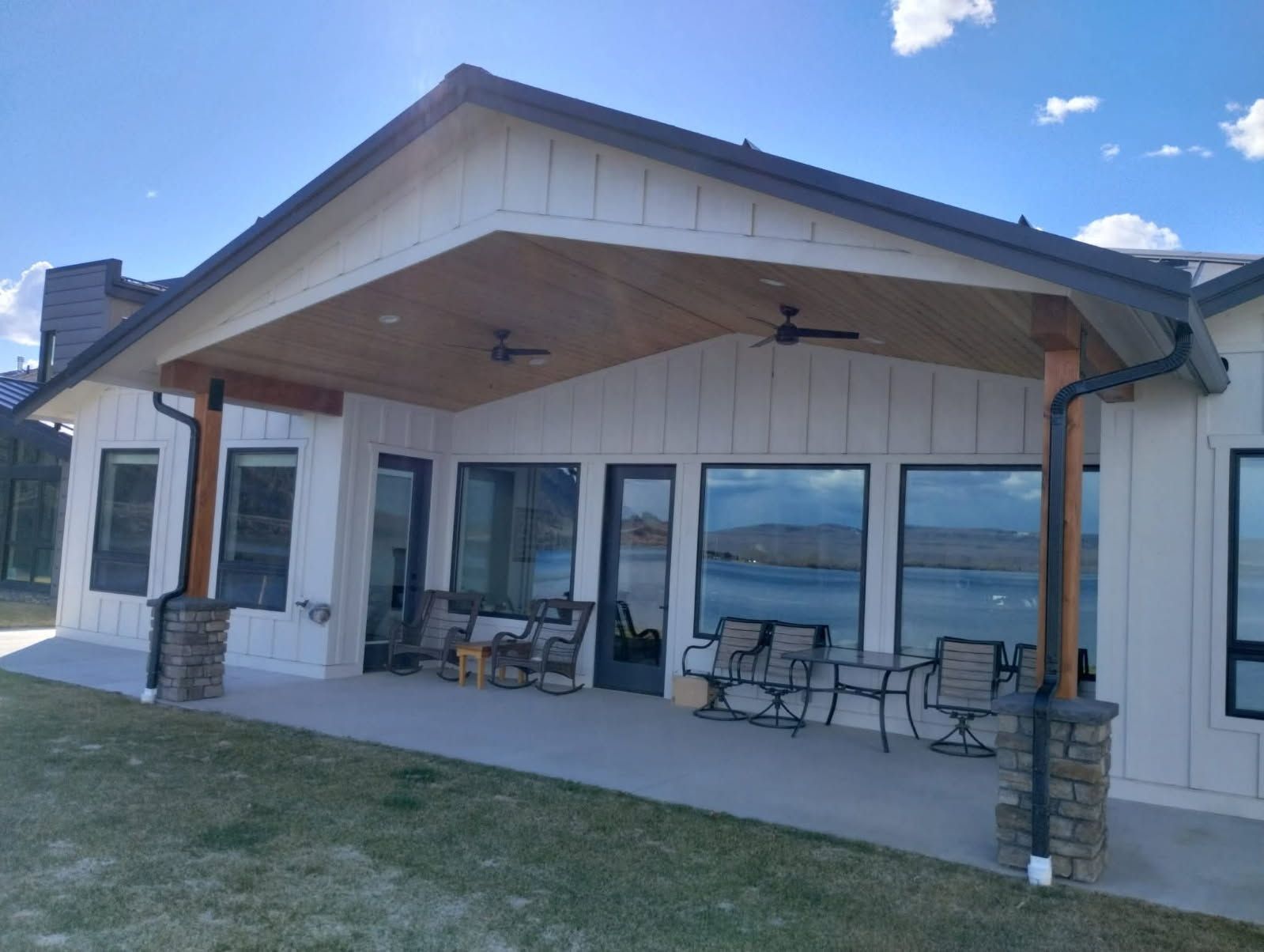 A modern, white farmhouse with a large covered patio and stone-based pillars, overlooking a lake under a sunny sky.