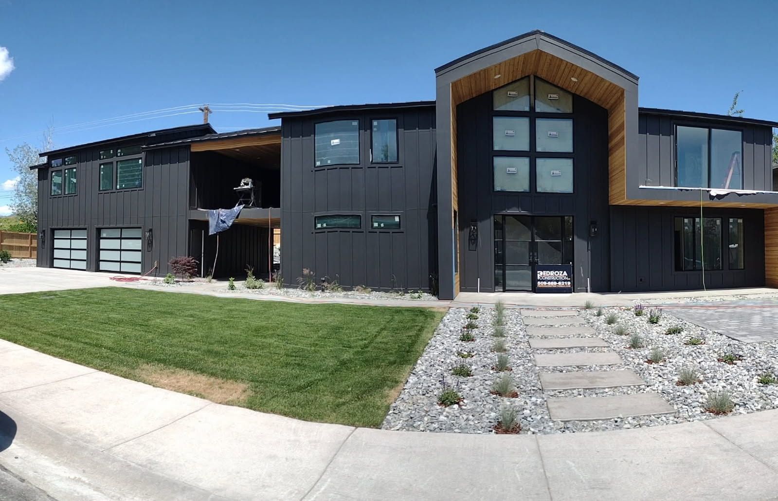 Modern two-story black house with wood accents, a wide garage, green lawn, and a stone walkway under a clear blue sky.