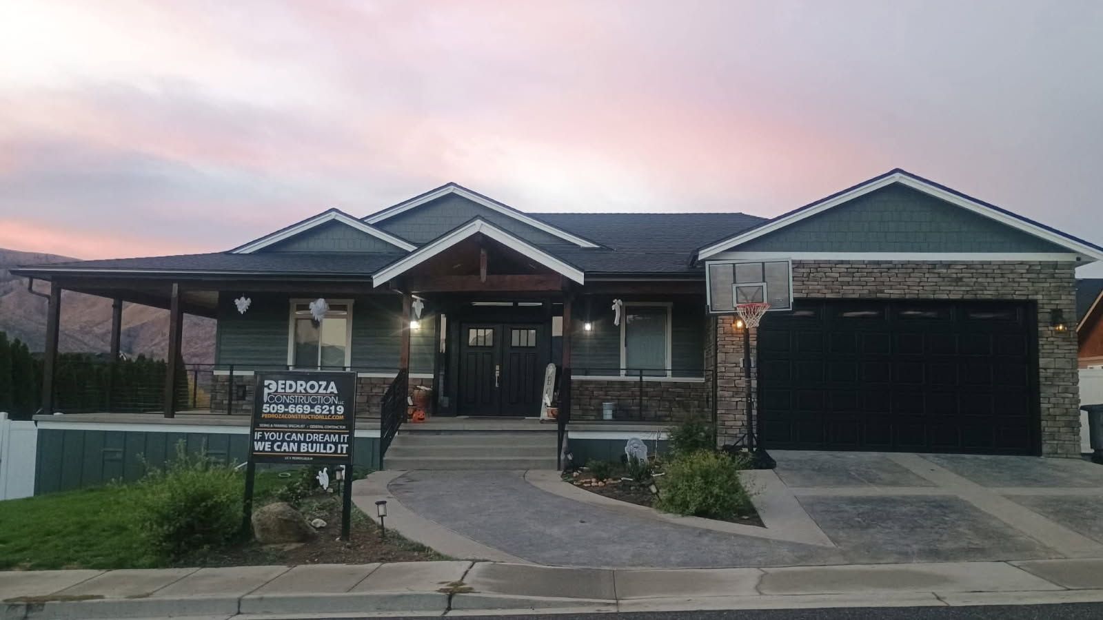 Single-story house with dark blue siding, stone accents, a garage, and a front porch at sunset under a pink sky.