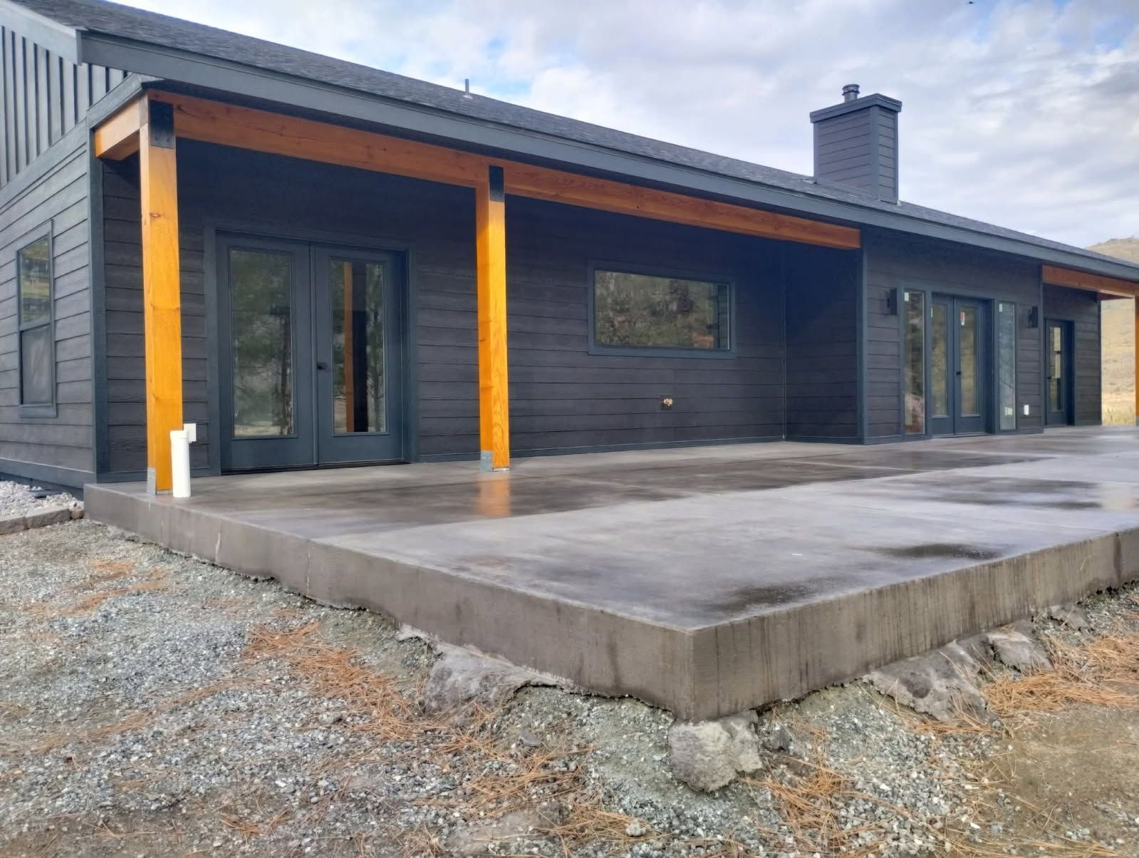 Modern one-story house with dark siding, a large concrete patio, and natural wood support beams under a roof overhang.