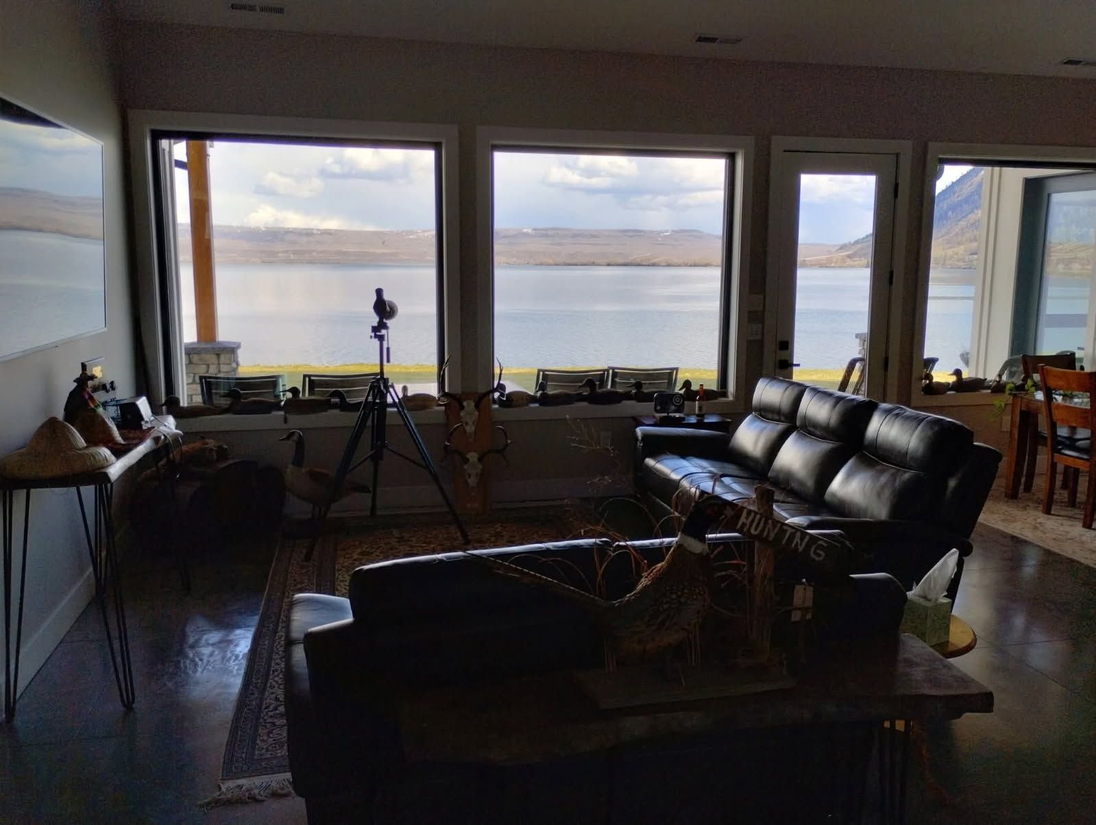 Living room with a black leather sofa, a tripod facing large windows, and a scenic view of a lake and hills.