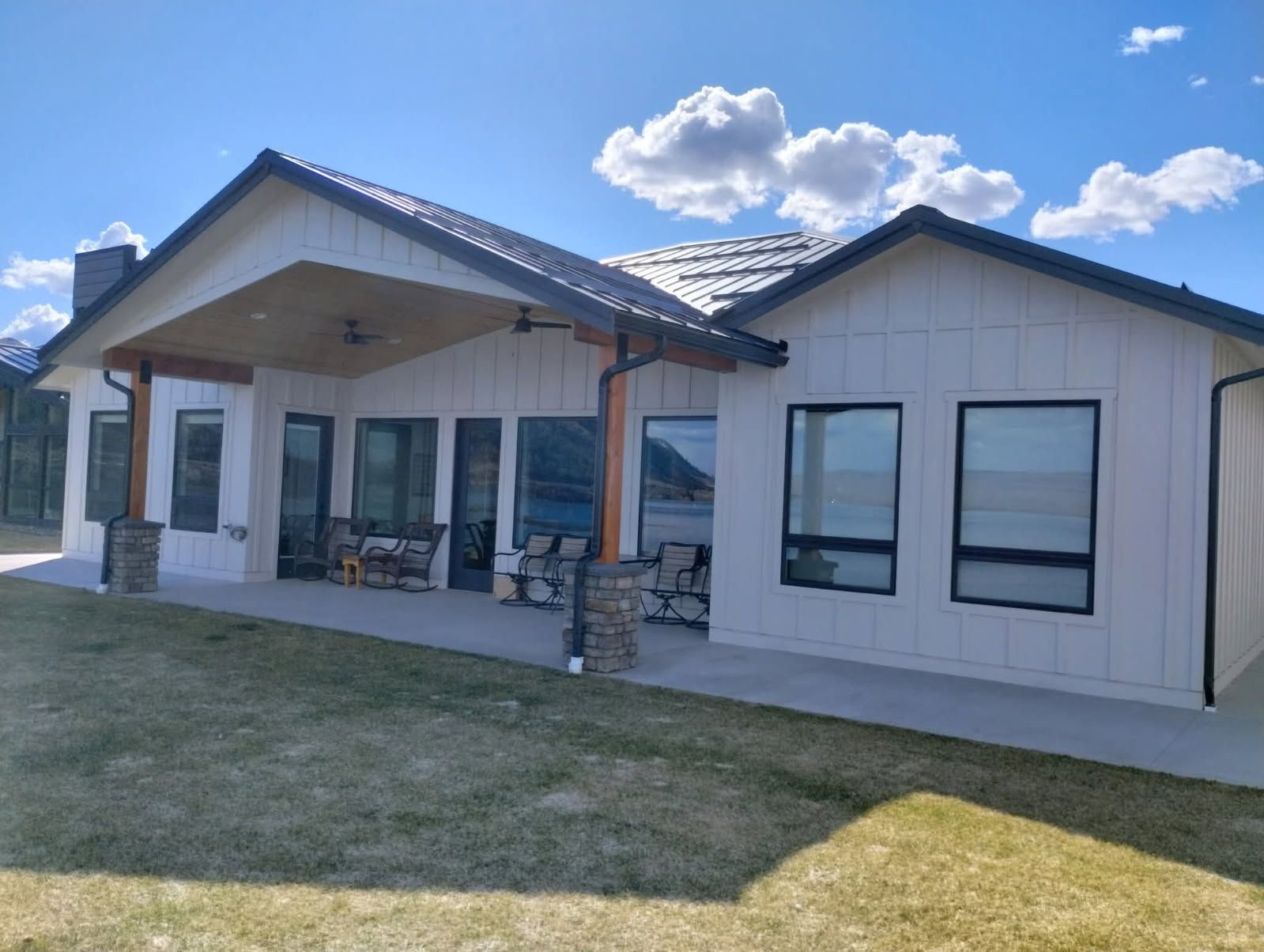Modern white farmhouse with black trim and roof, featuring a covered patio and a large lawn under a blue sky.