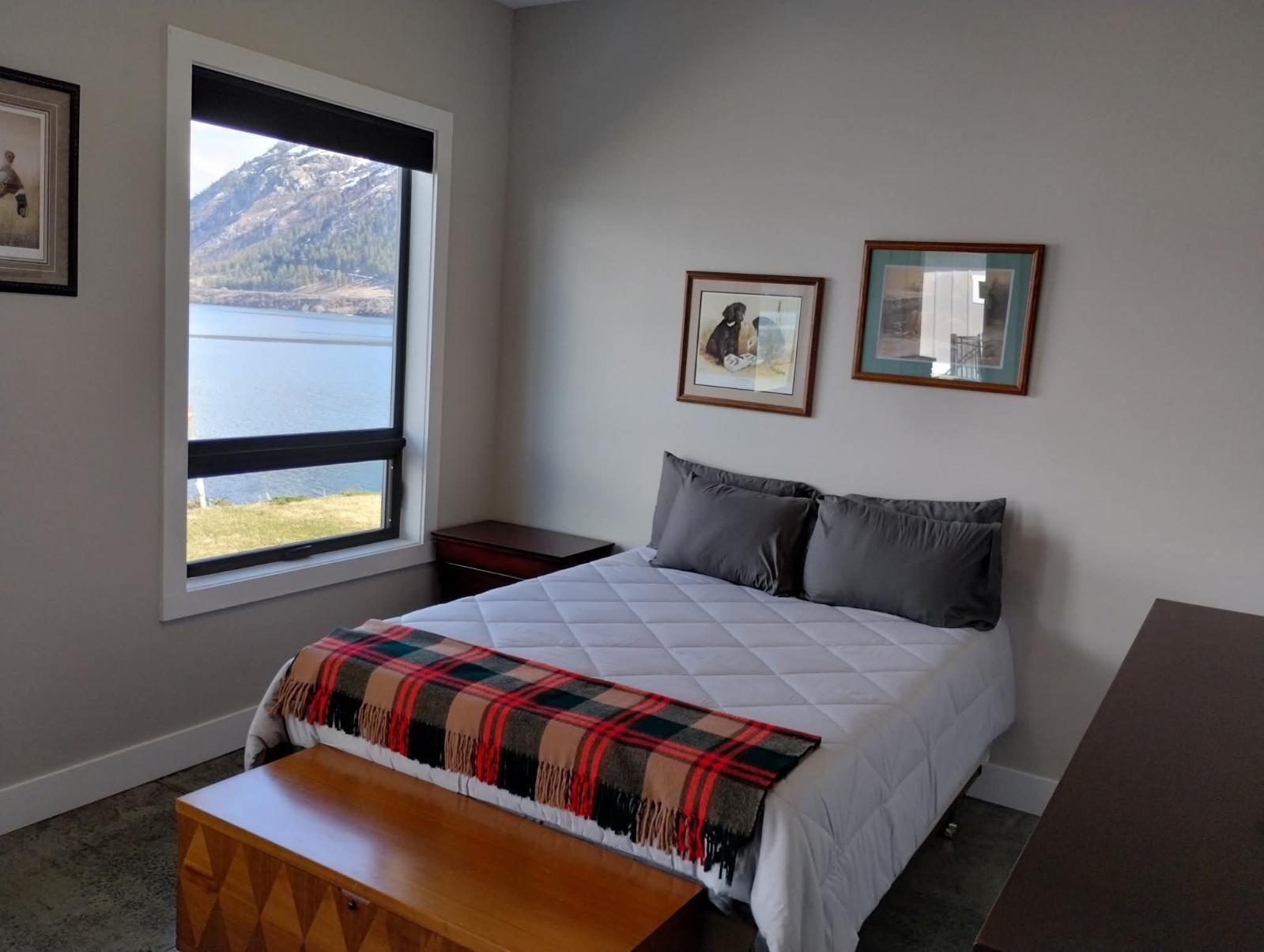 A bedroom with a queen bed featuring a white quilt and plaid throw, wood chest, and a window with a mountain lake view.
