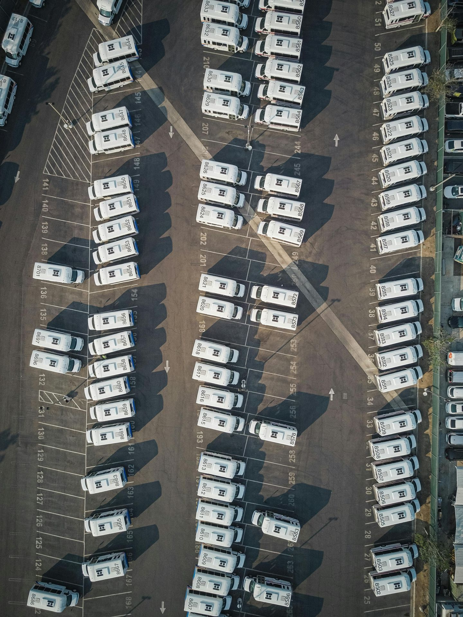 Luchtfoto van rijen witte auto's geparkeerd op een parkeerterrein met smalle rijstroken ertussen.
