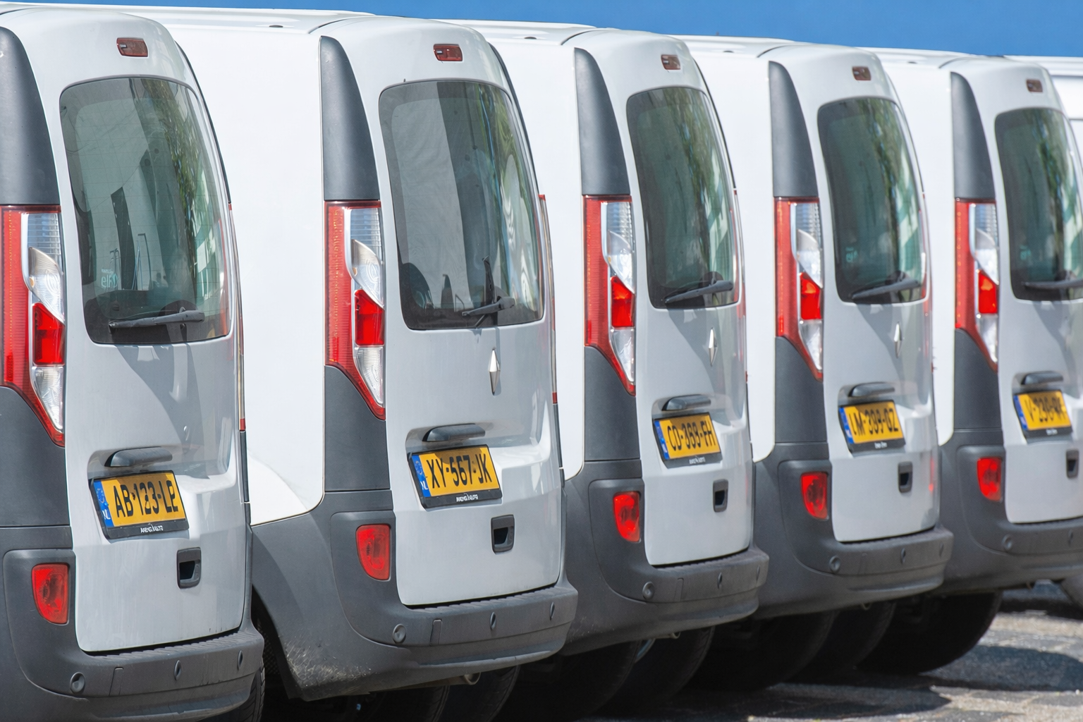 Achteraanzicht van een aantal zilverkleurige bestelbusjes die op een rij geparkeerd staan met gele kentekenplaten.