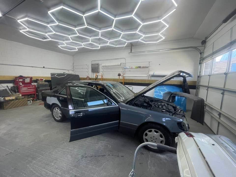 Car in a garage with hood open and driver door ajar under bright hexagon ceiling lights