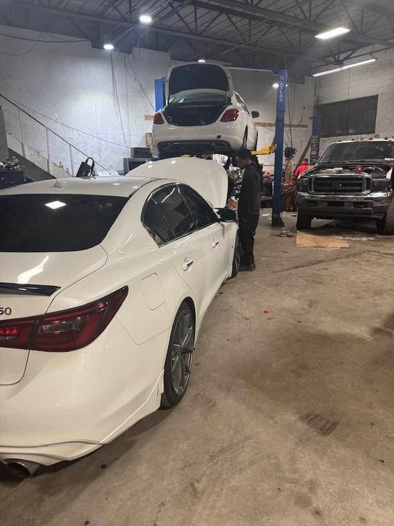 White sedan in a repair shop with its trunk open, parked beside a black SUV and another car on a lift.