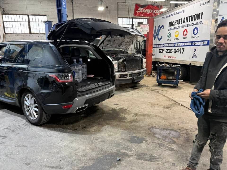 SUV with open hatch in auto repair shop, mechanic working nearby, service truck parked beside it