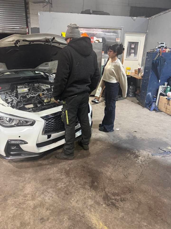 Two people inspect a white car with its hood open in an auto repair shop.
