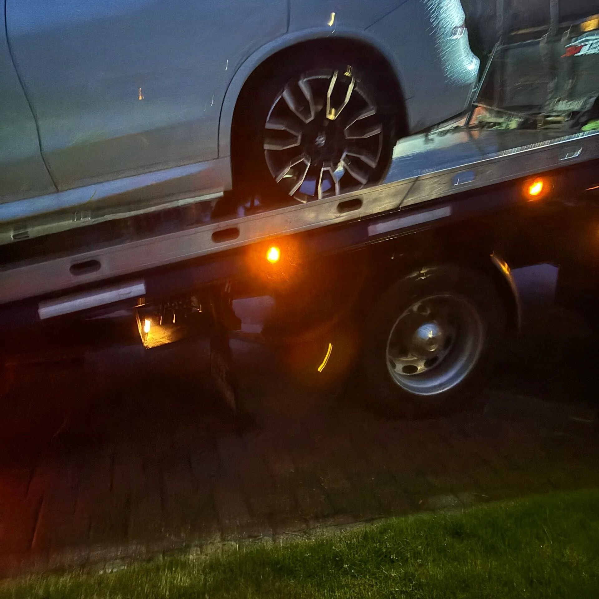 Silver car on a tow truck at night, with orange warning lights glowing.