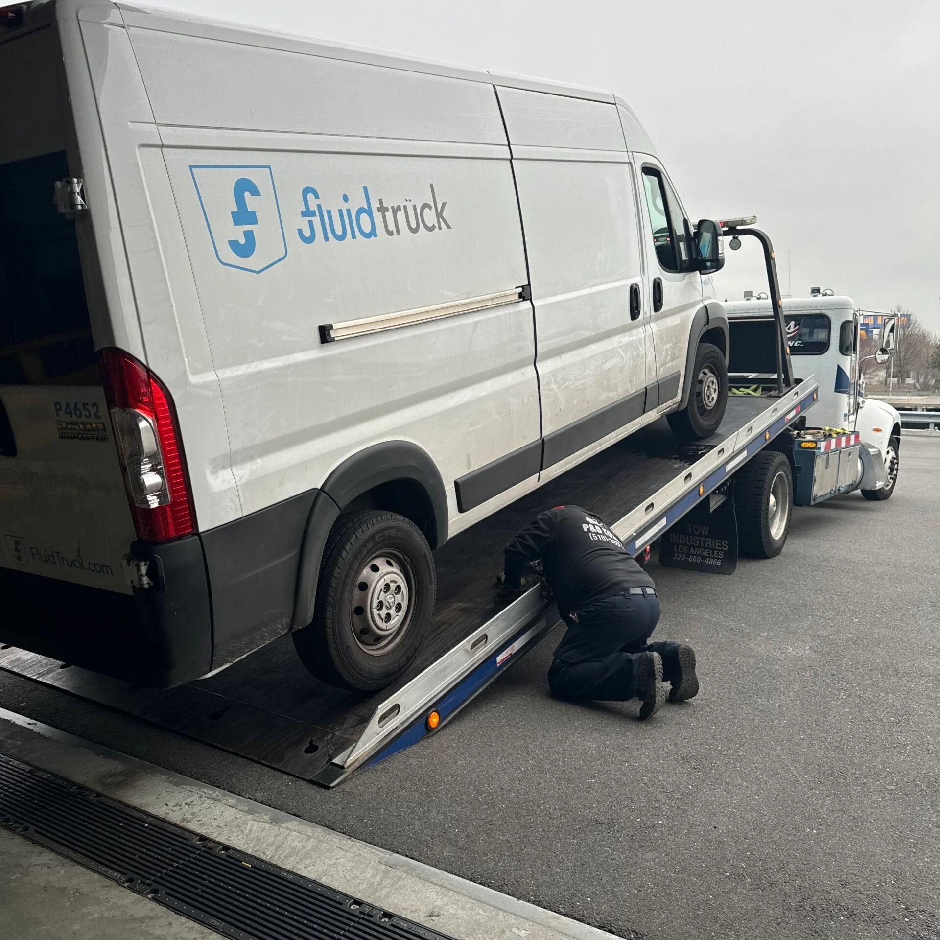 White van being loaded onto a flatbed tow truck on a roadside, with a worker kneeling beside it