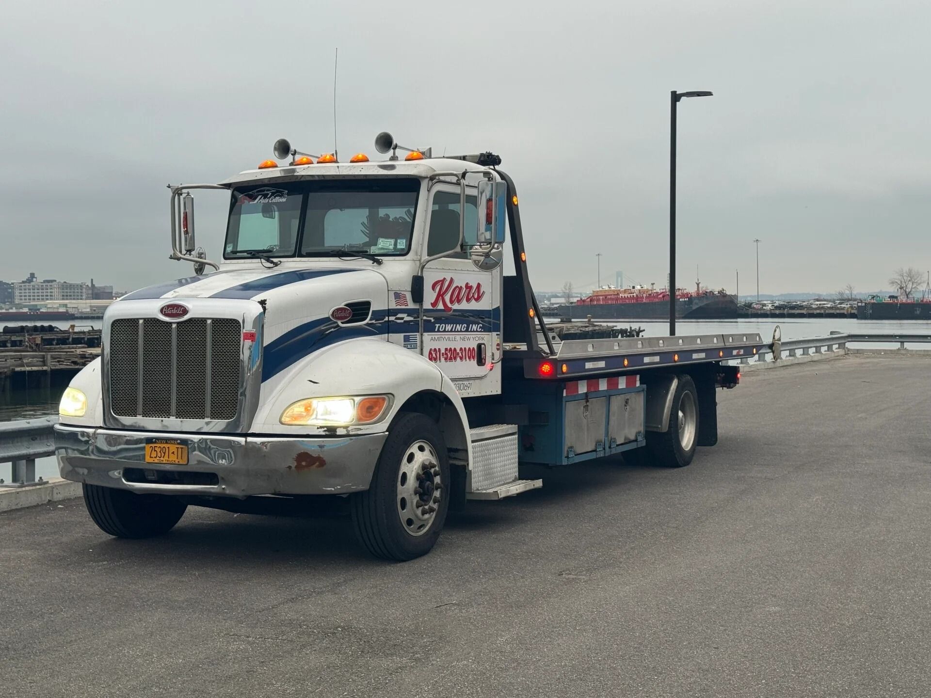 Tow truck parked on a snowy lot, with headlights on and a flatbed behind a white cab.