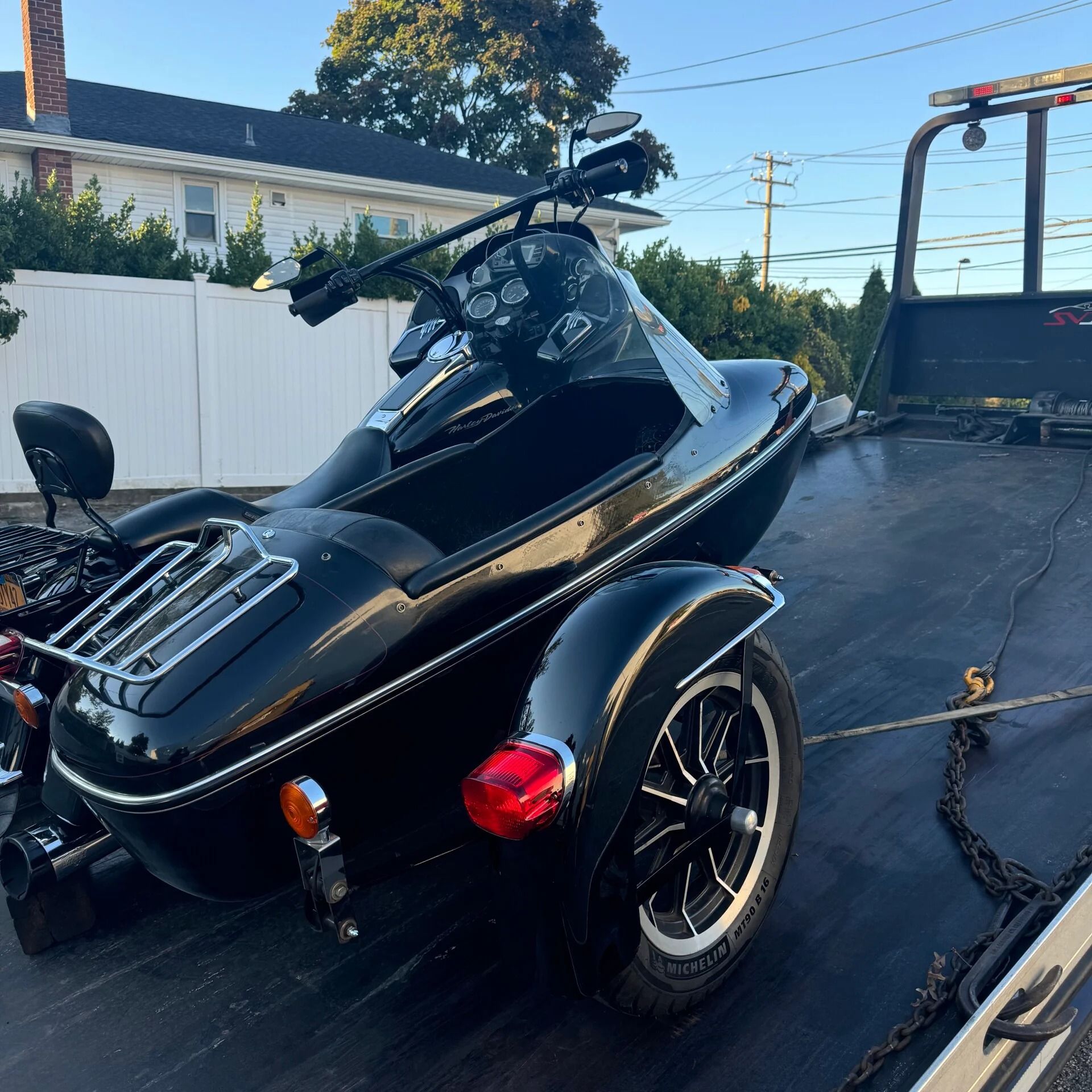 Black three-wheeled motorcycle on a trailer in a driveway, with a white fence and blue sky in the background