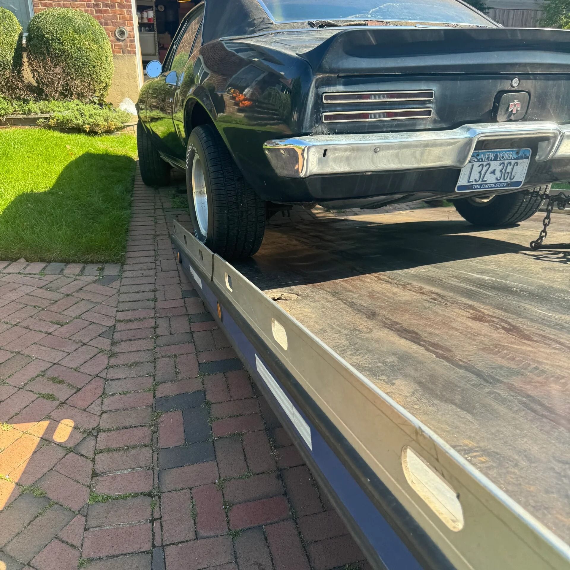 Black classic car on a tow truck next to a brick sidewalk in a sunny residential area