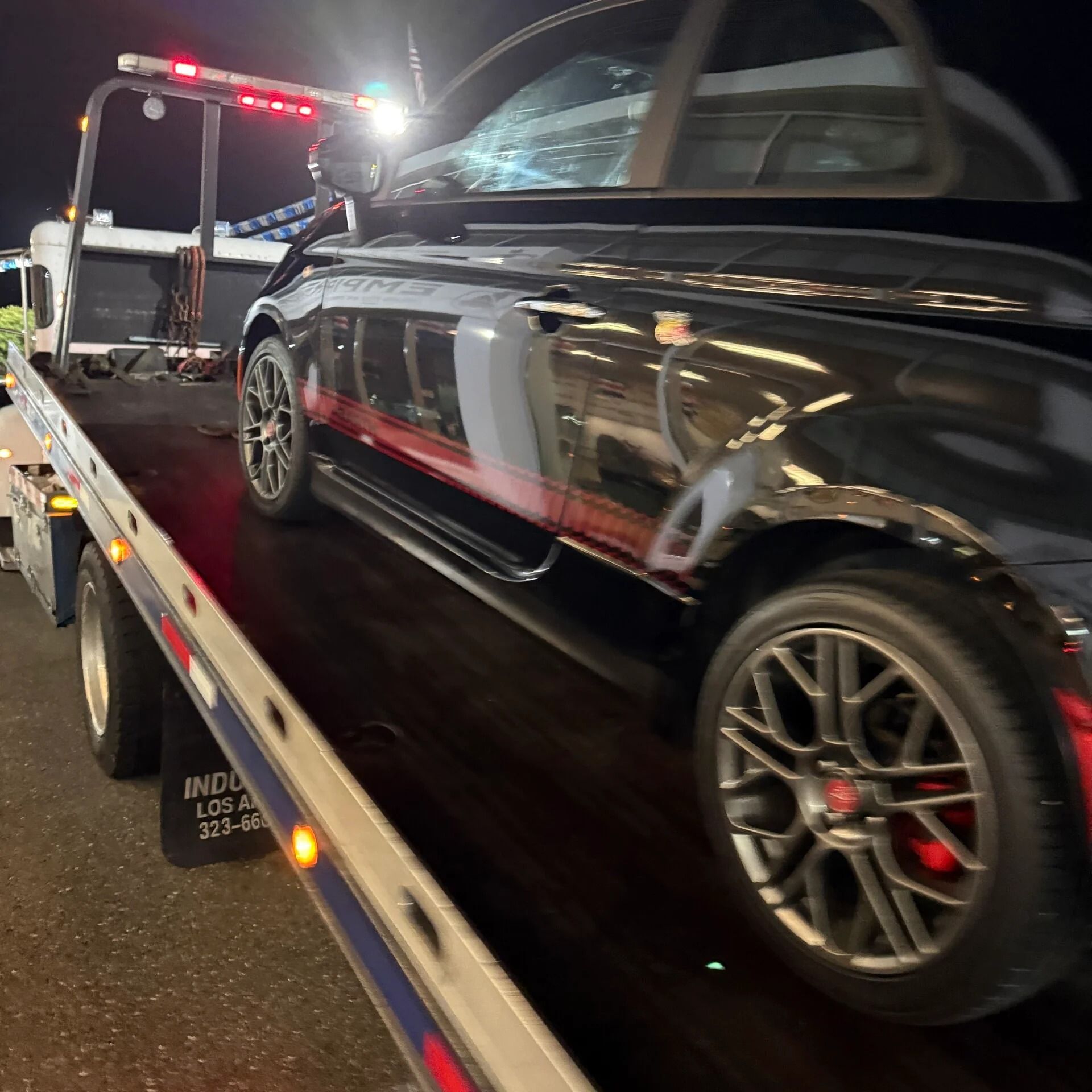 Black convertible being loaded onto a tow truck at night, with red warning lights visible.