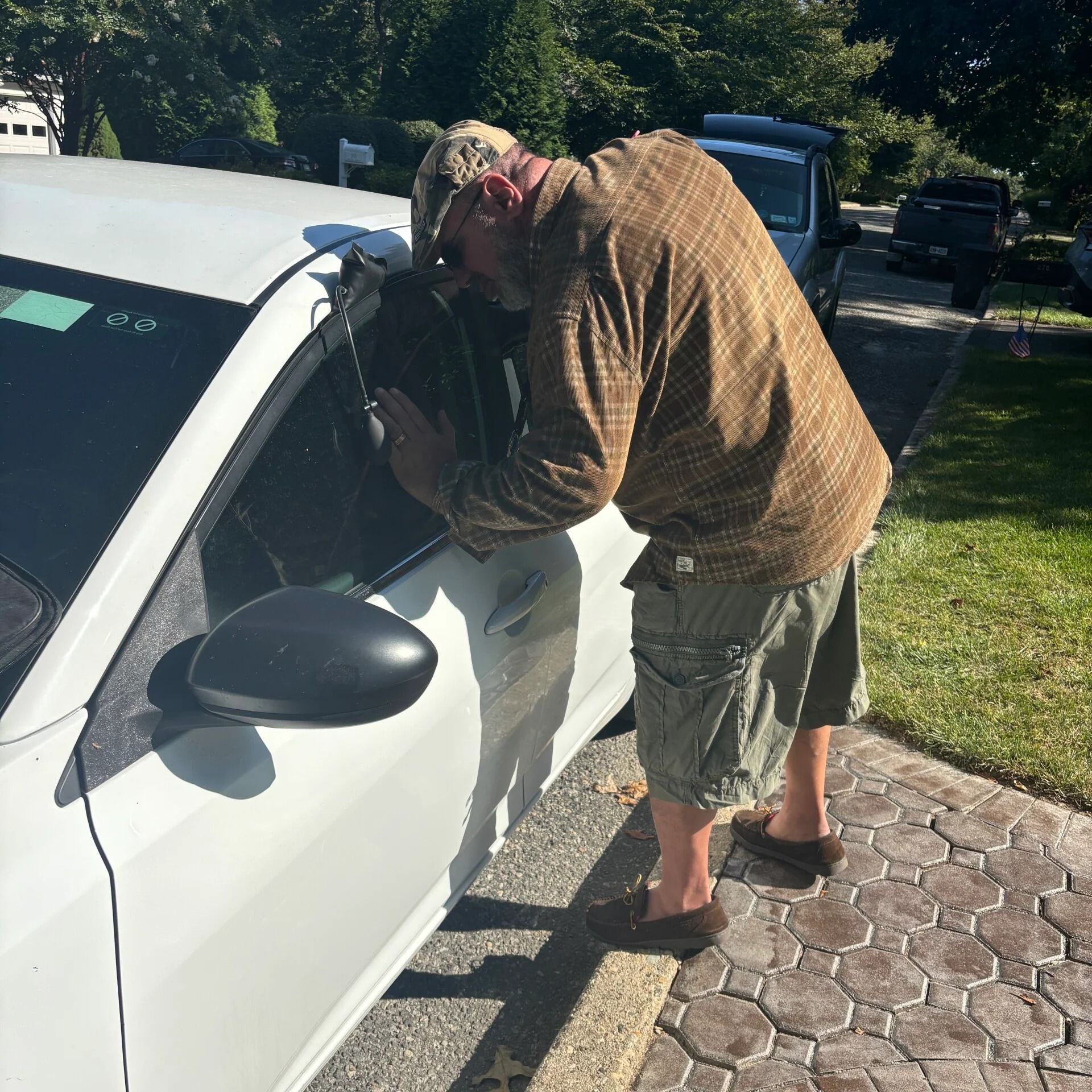 Person leans into a white car’s open window while another stands nearby on a sunny driveway.