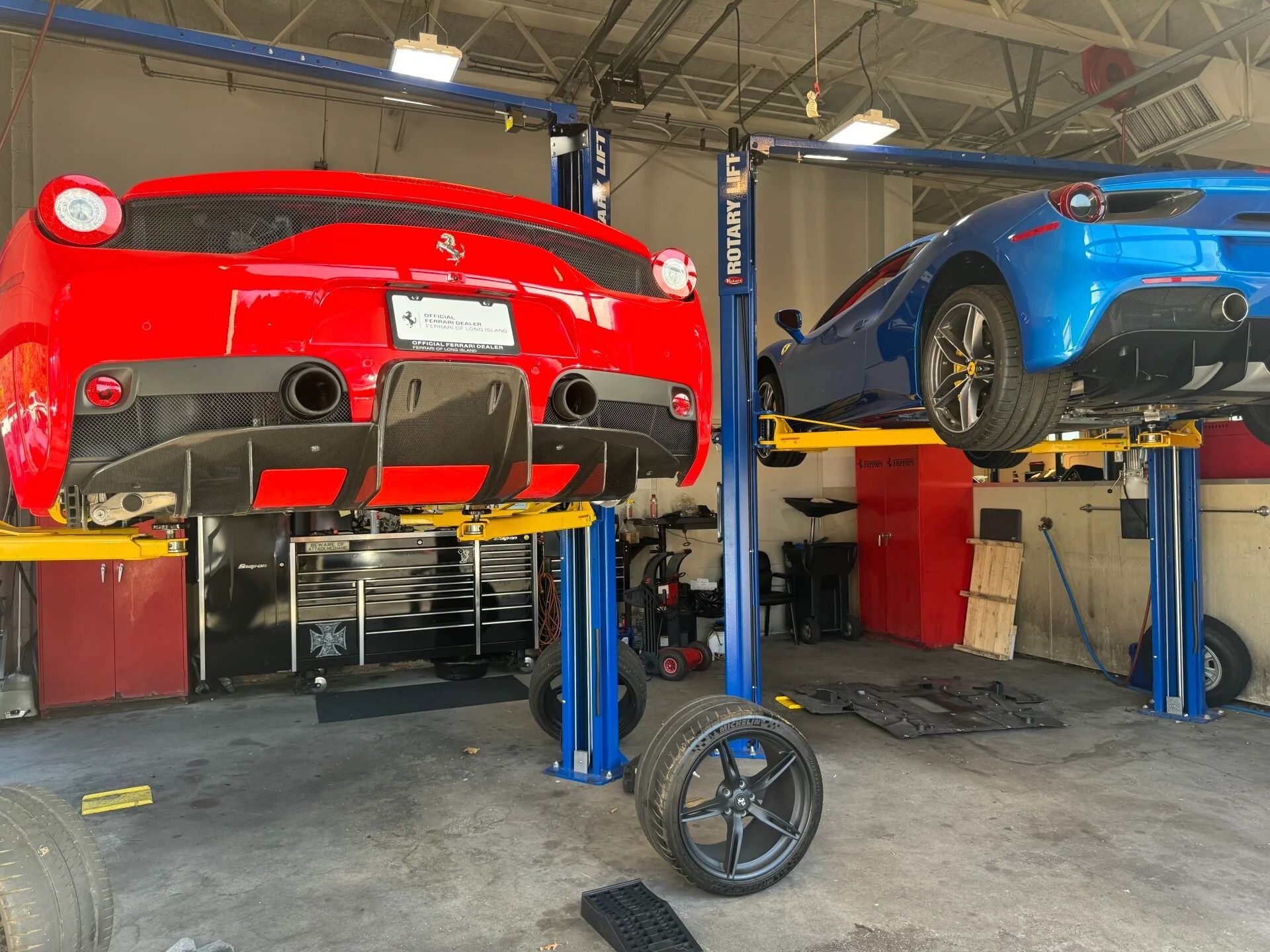 Red sports car and blue car lifted in an auto repair garage, with a wheel on the floor.