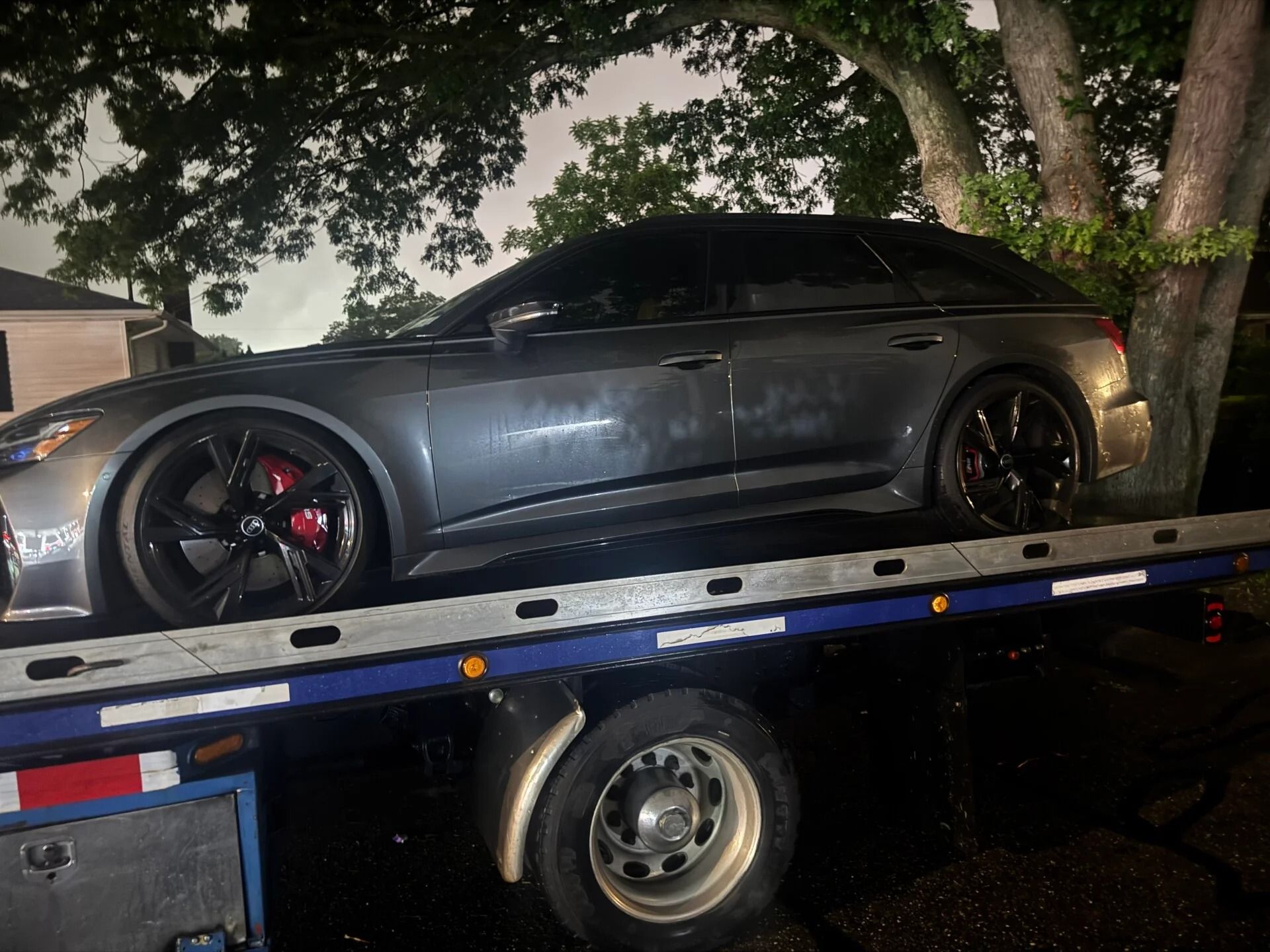Dark gray sports coupe on a tow truck at night, parked near trees and houses