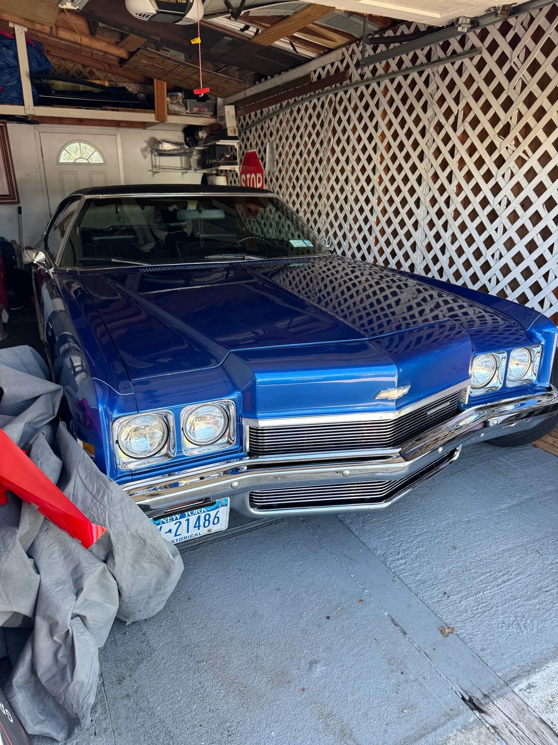 Blue classic car parked in a carport with a lattice wall and overhead storage bins