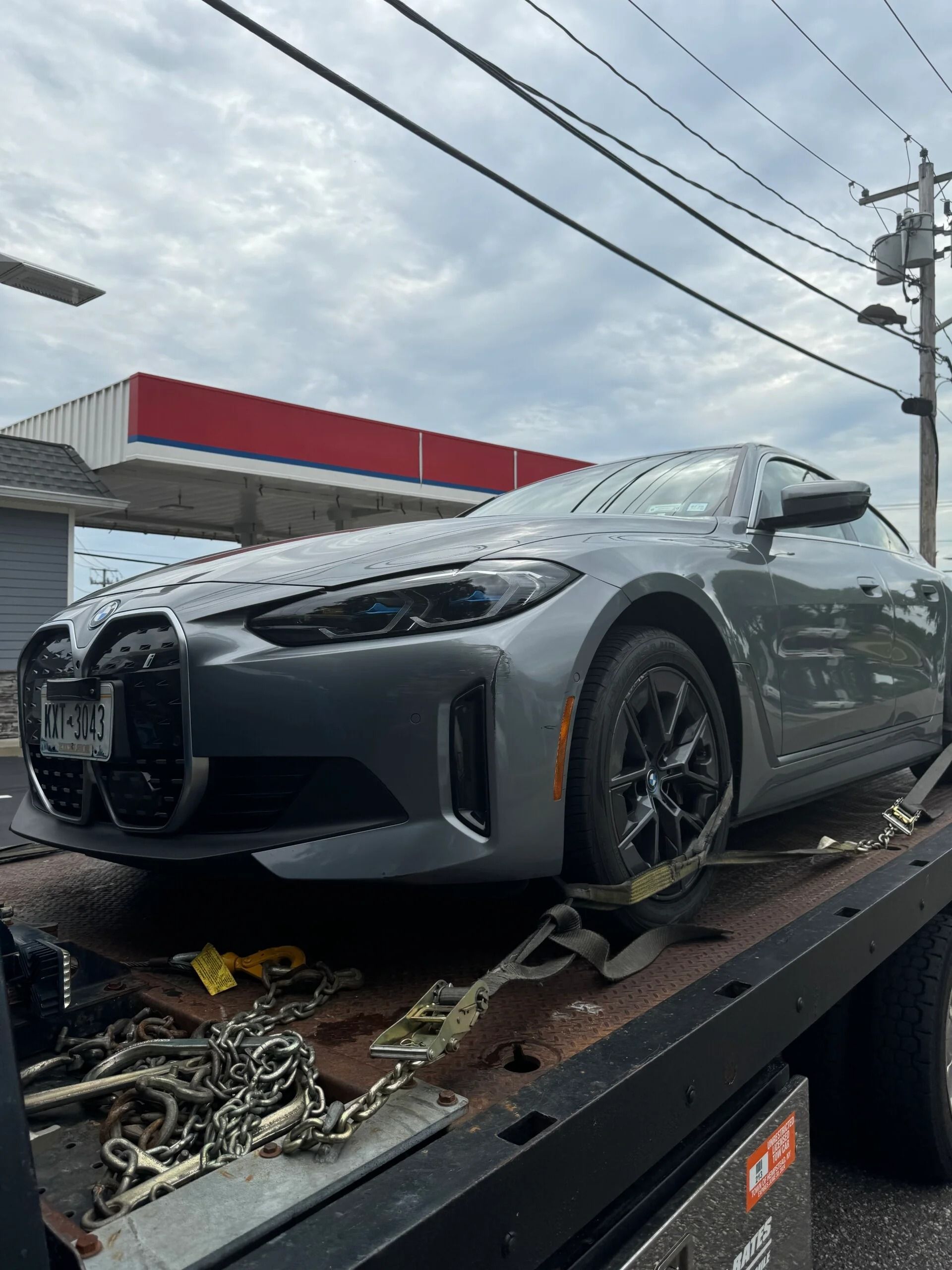 Gray sports car on a tow truck bed, front damage visible under a cloudy sky.
