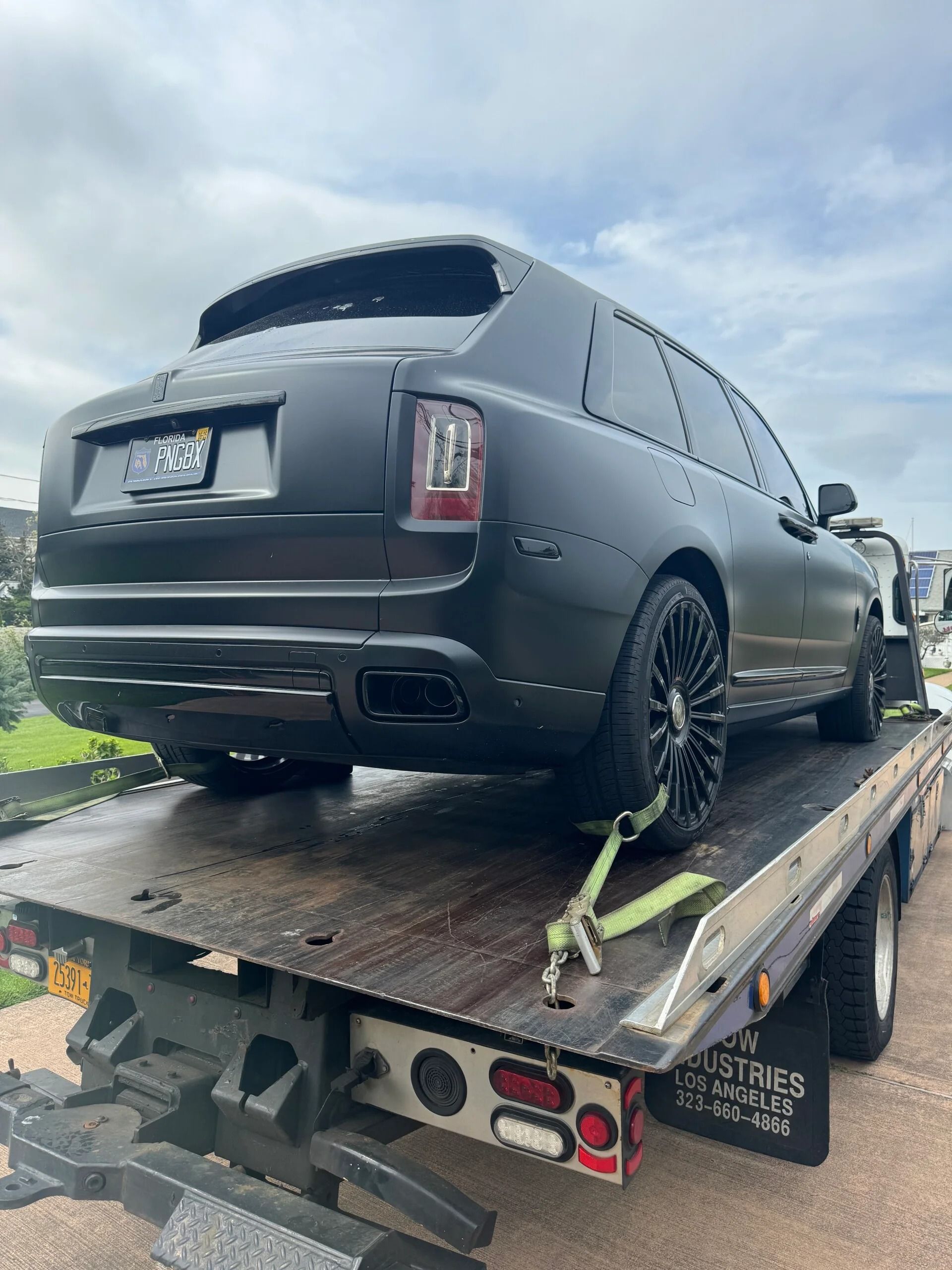 Black SUV loaded on a tow truck flatbed, secured with straps on a wet road under cloudy skies