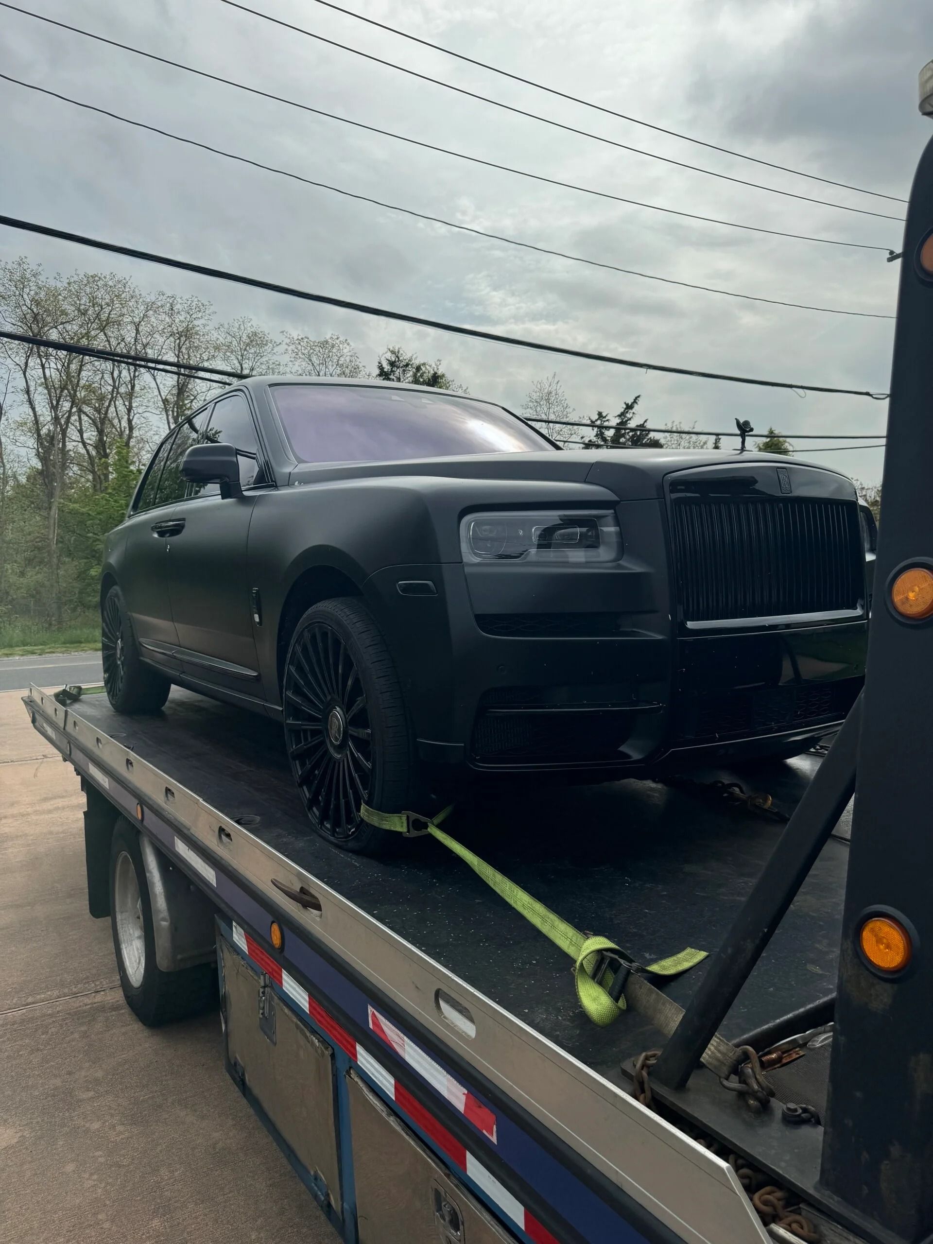Matte black Rolls-Royce SUV strapped on a flatbed tow truck under cloudy skies