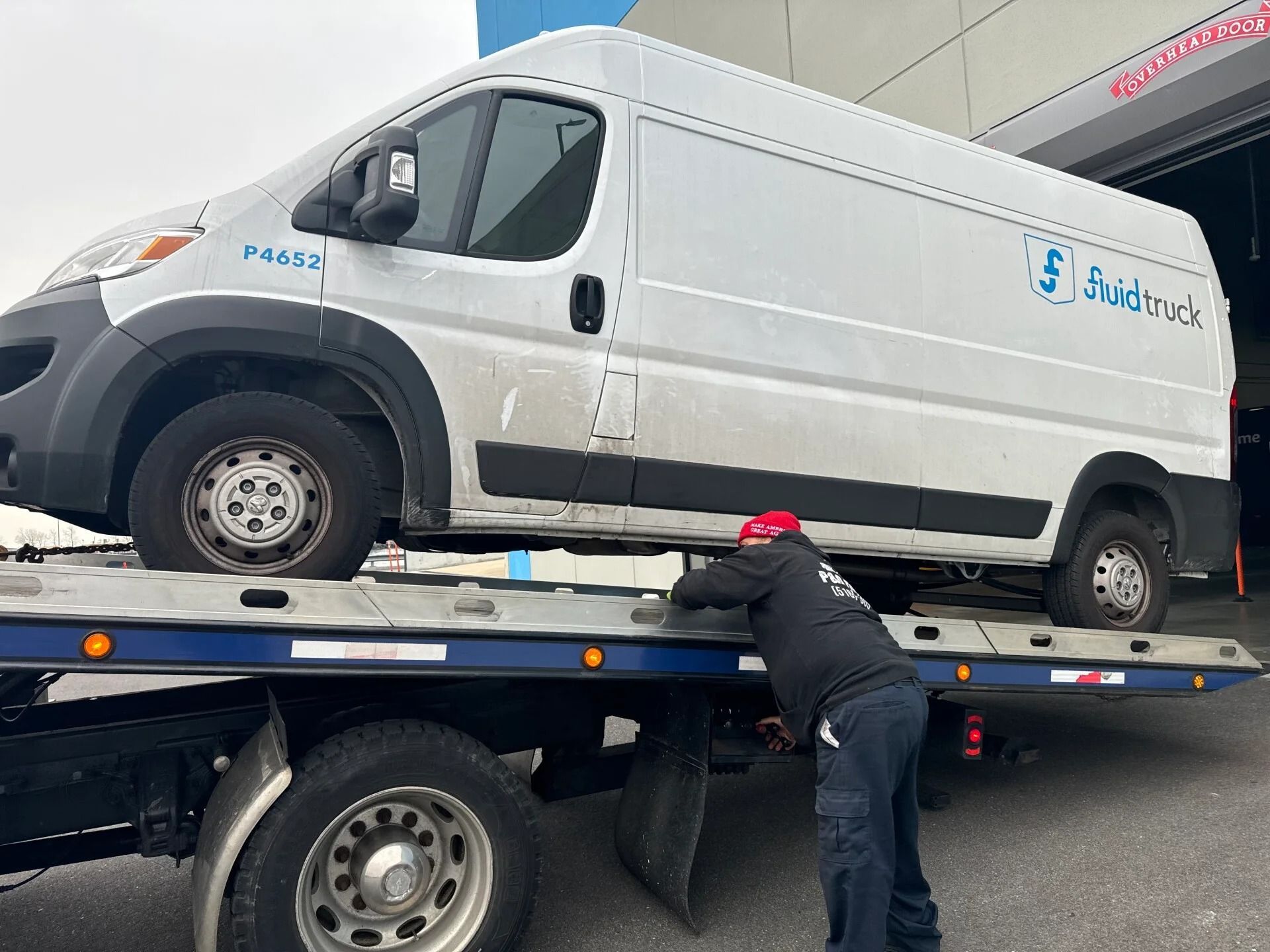 Worker securing a white van on a flatbed tow truck with a chain in a parking lot