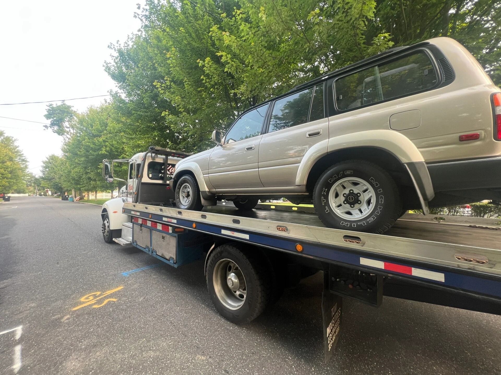 Flatbed tow truck carrying a beige SUV on a tree-lined road