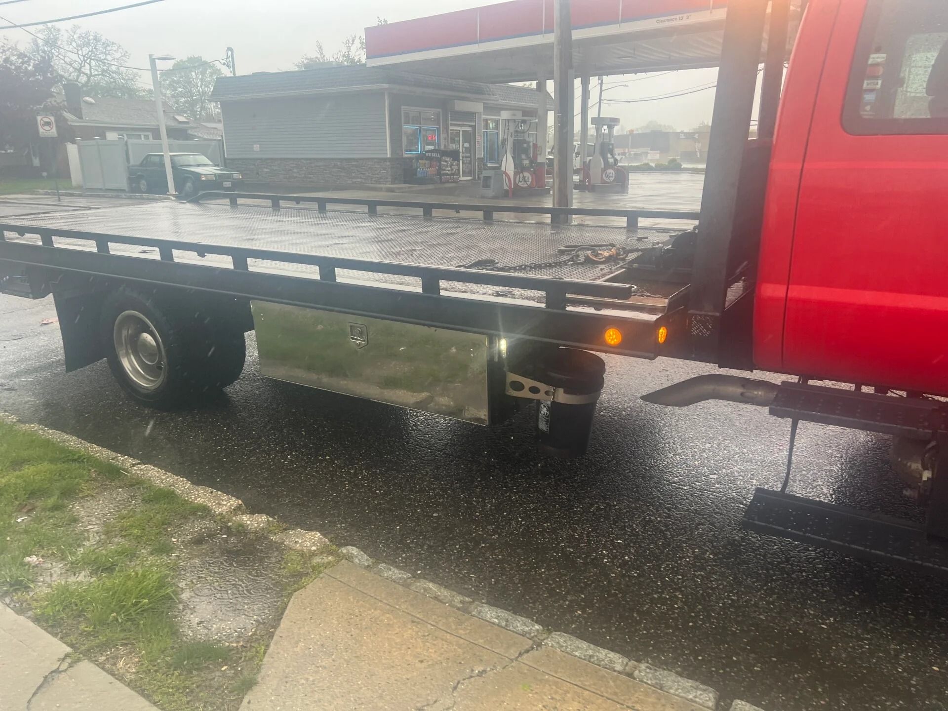 Red flatbed tow truck parked on a wet street near a gas station, rear lights on.