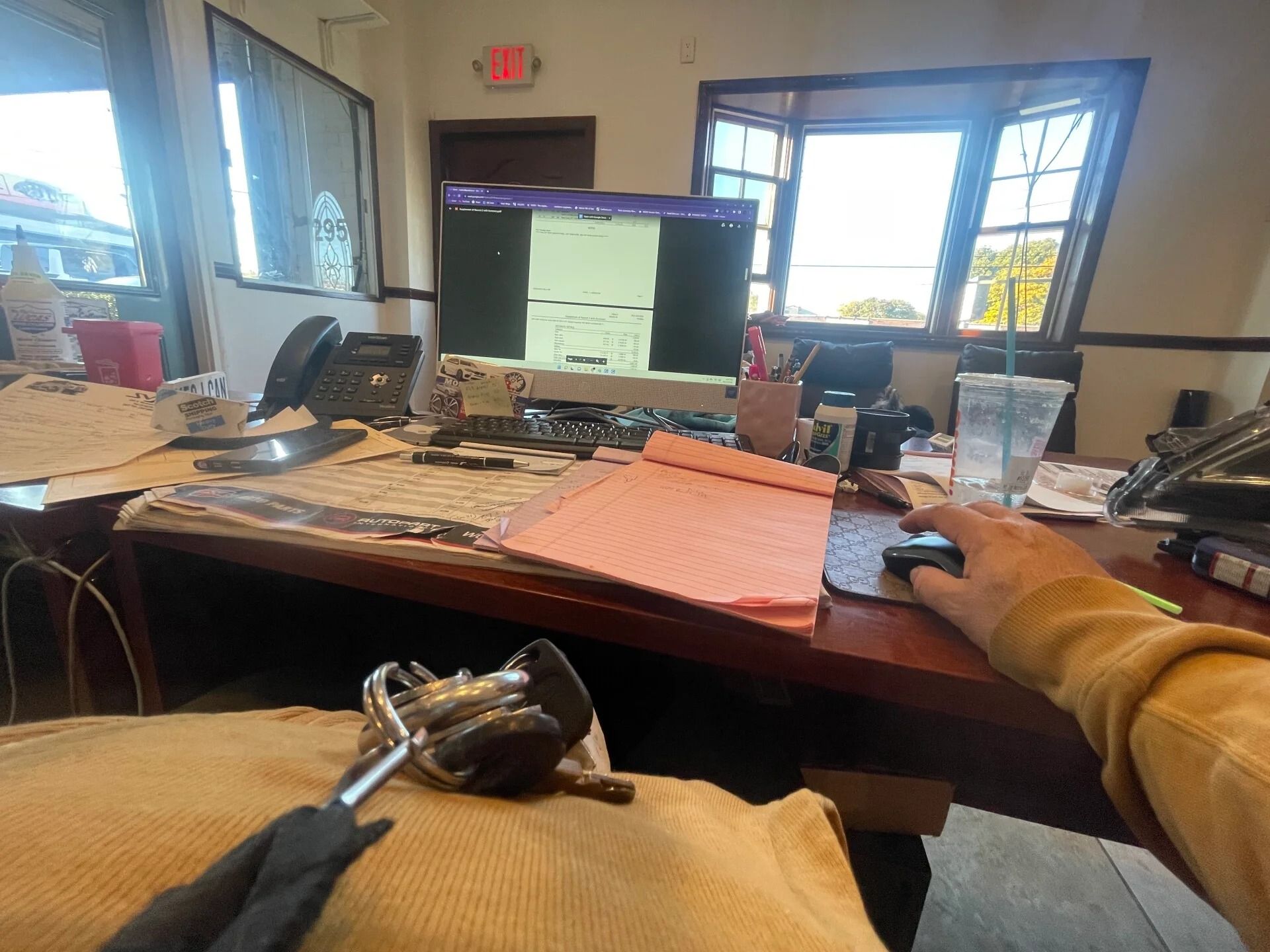 Office desk with computer monitor, papers, keys, and a person’s arm using a mouse near a window