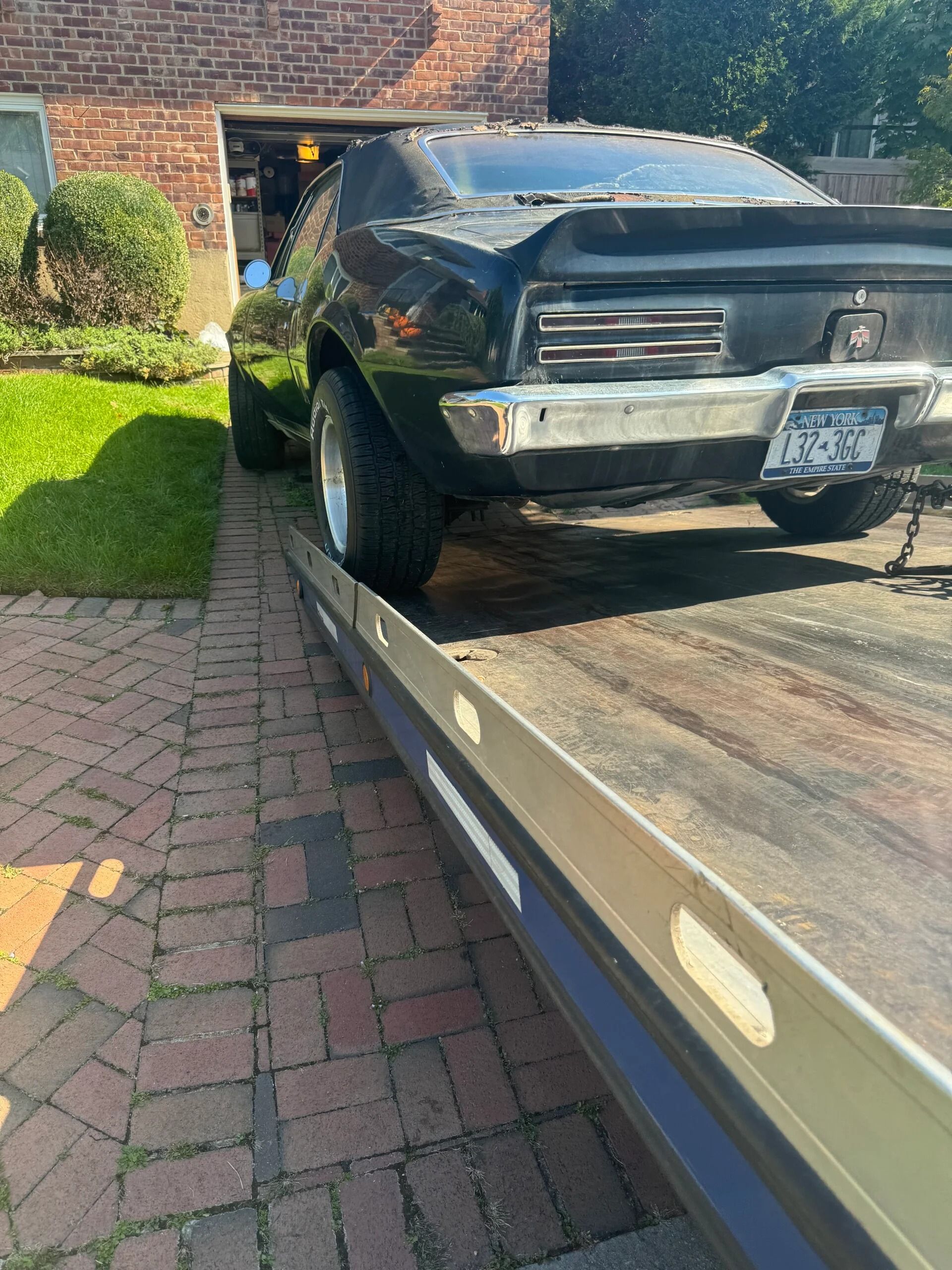 Black classic car partly on a flatbed tow truck in a brick driveway beside a garage.